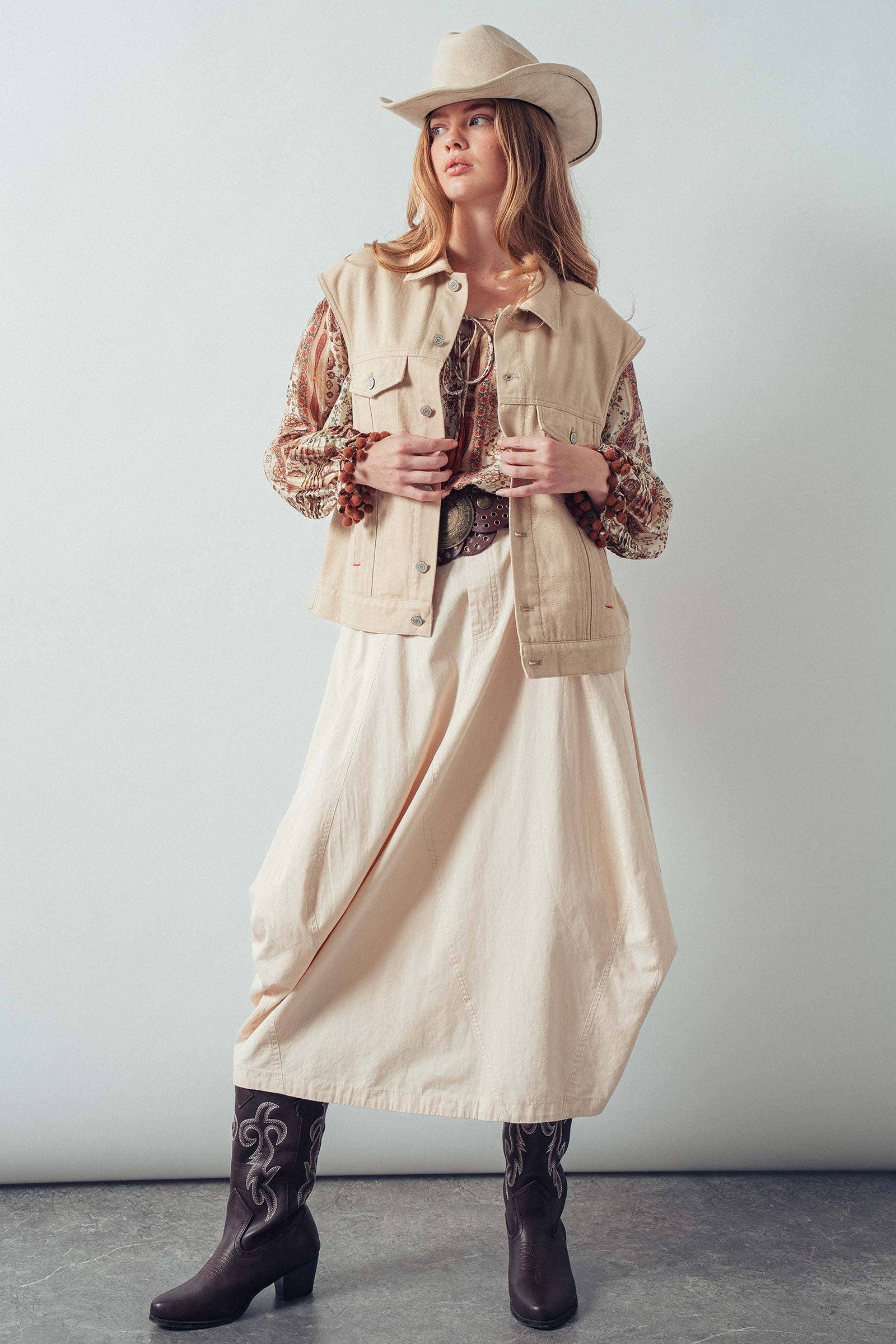 Woman wearing a beige vest, long skirt, and cowboy hat against a white background