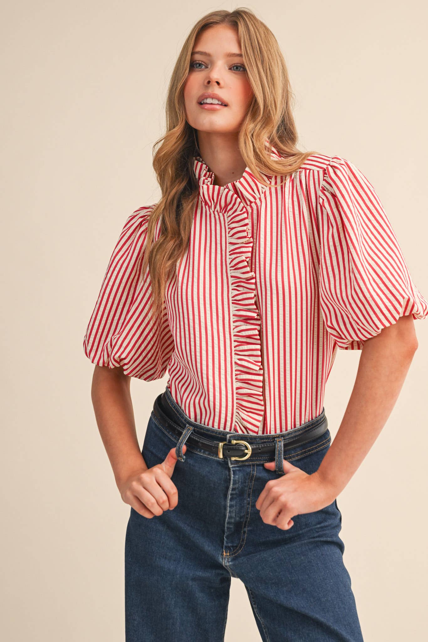 Woman wearing a red and white striped blouse with puffed sleeves and blue jeans on a beige background