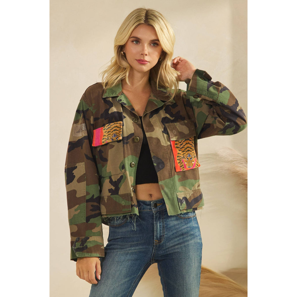 Woman wearing a camouflage jacket with colorful patches on a neutral background. Styled with crop tank and denim jeans.