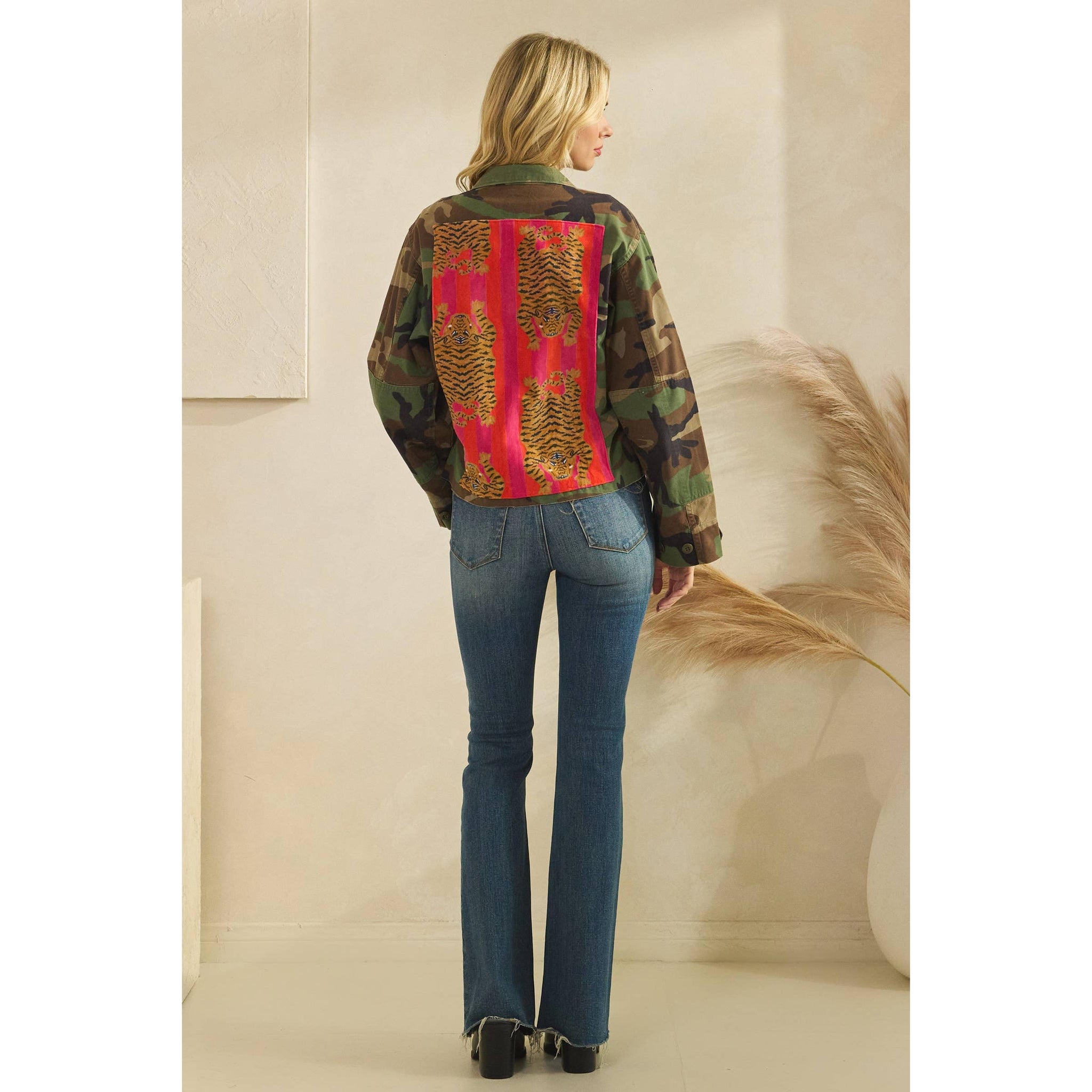 Woman wearing a camouflage jacket with a red and yellow tiger pattern on the back, standing against a neutral background. Styled with jeans and black boot. Full length back view.