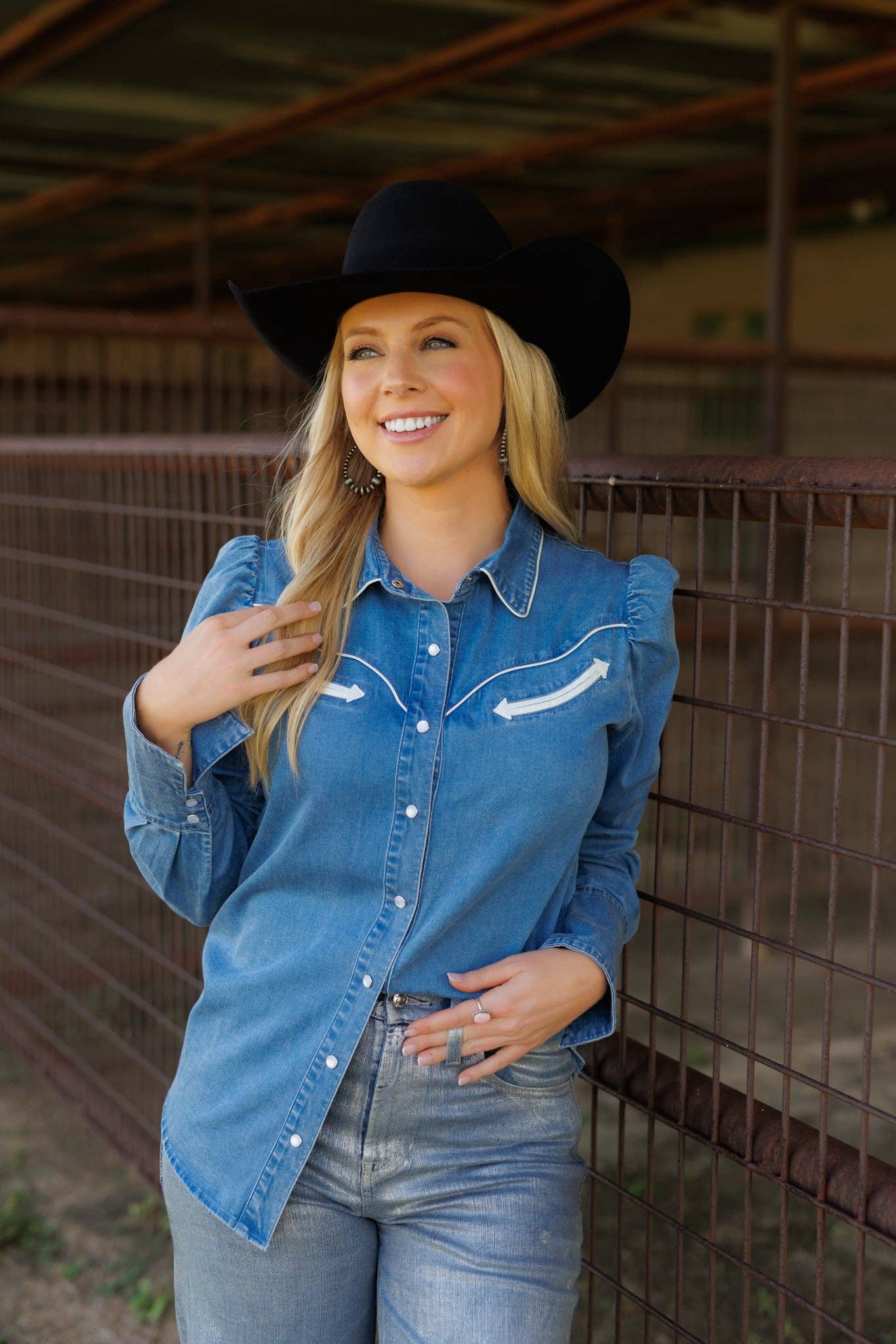 Woman wearing a blue denim shirt with white arrow and a black cowboy hat, standing in an indoor setting.