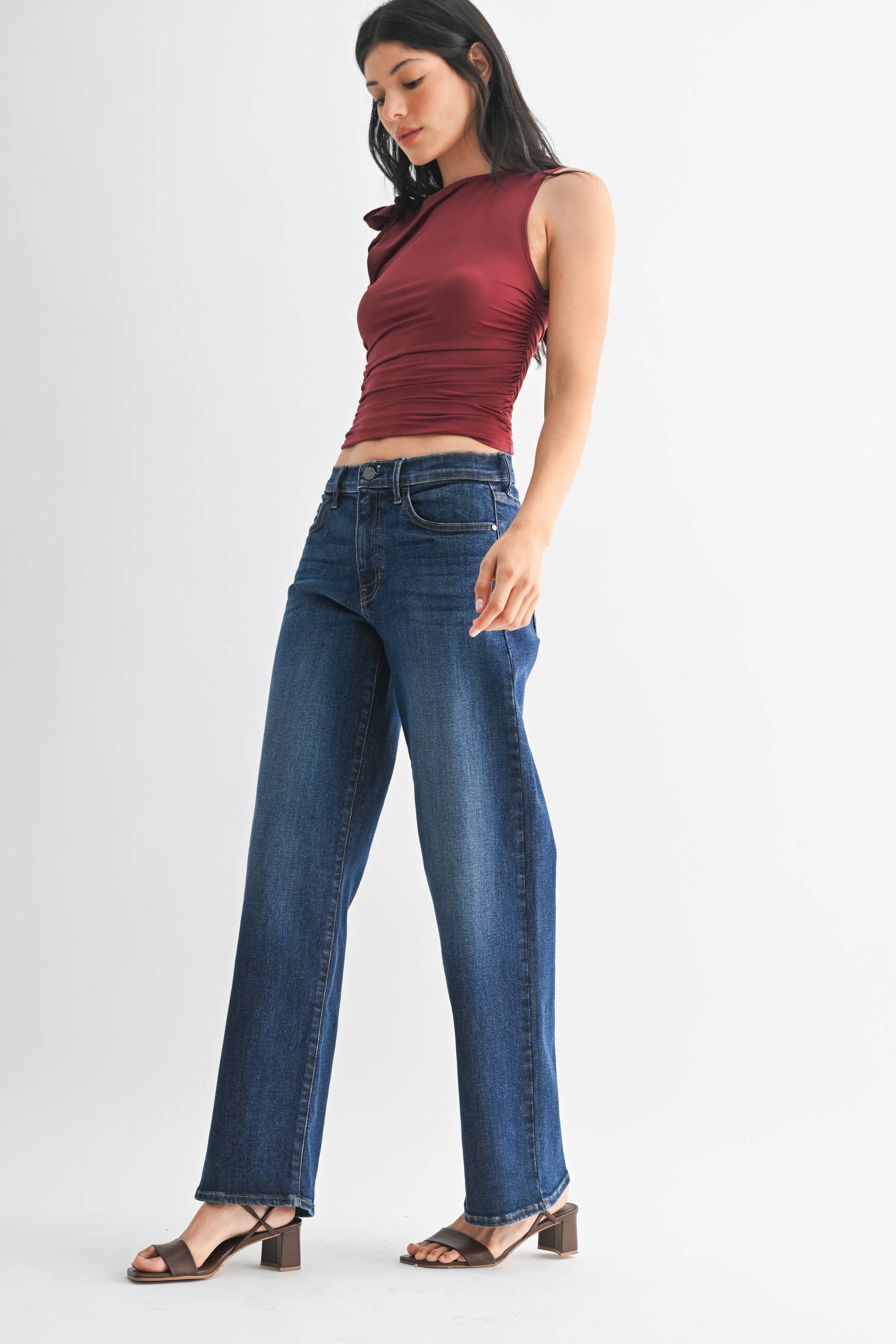 Woman wearing a red sleeveless top and blue jeans on a white background. Side view showing relaxed pant leg.
