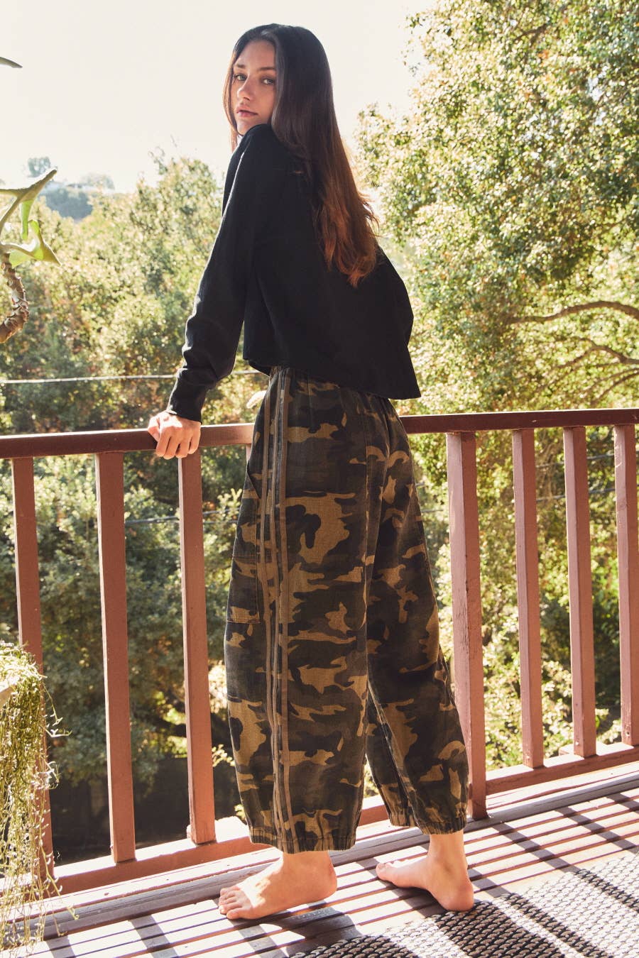Woman standing on a balcony wearing a black top and camouflage pants with trees in the background