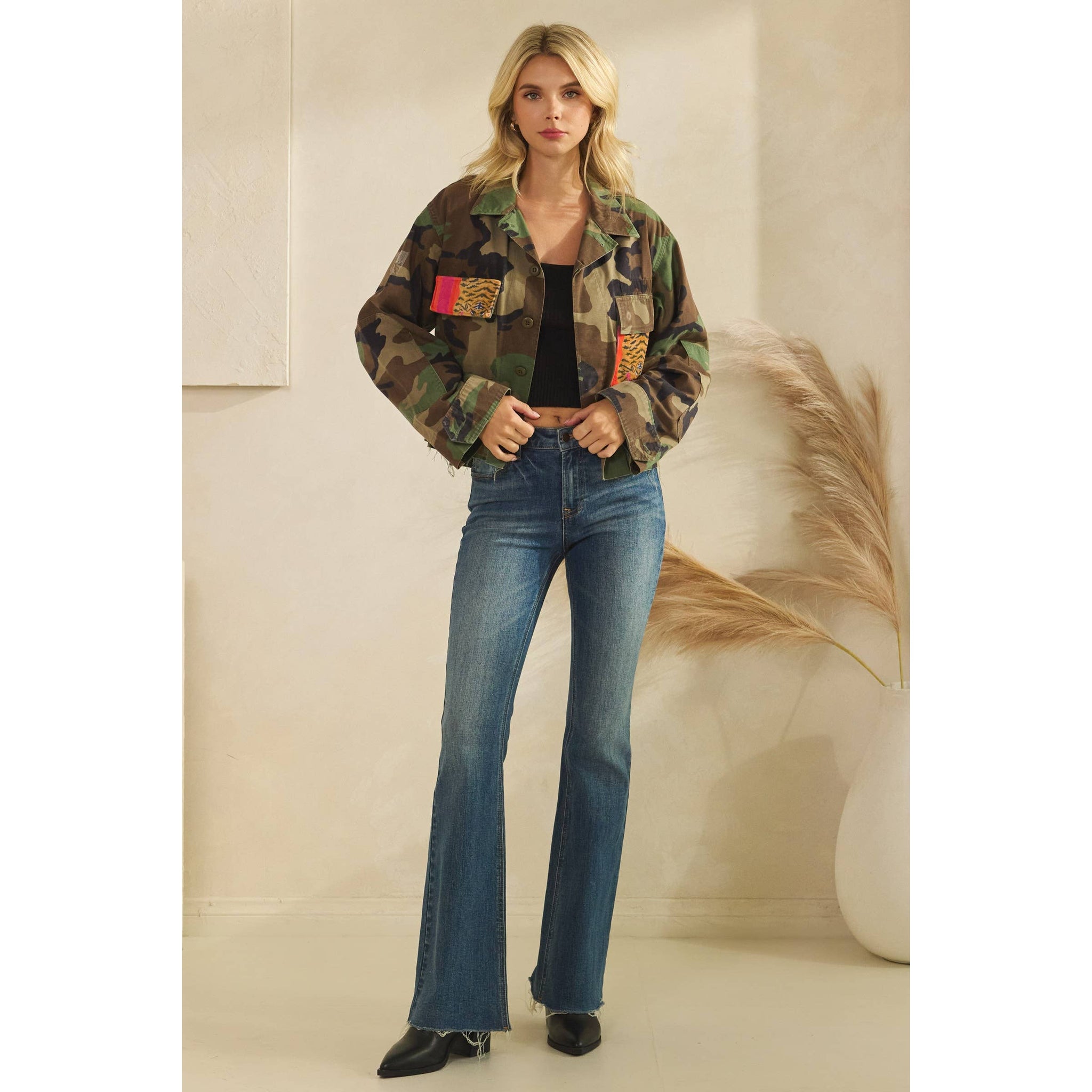 Woman wearing a camouflage jacket with colorful patches and blue jeans against a neutral background. Full length view.