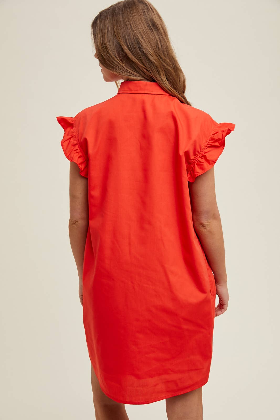 Woman wearing a red dress with ruffled sleeves on a beige background showing back view.