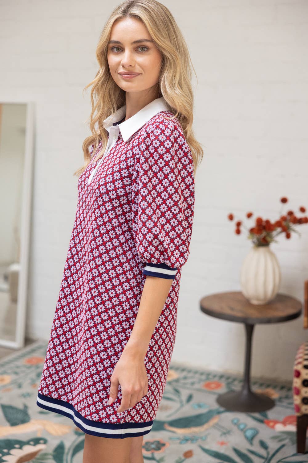 Woman wearing a red floral dress with white collar and blue trim in a room with a rug and table.