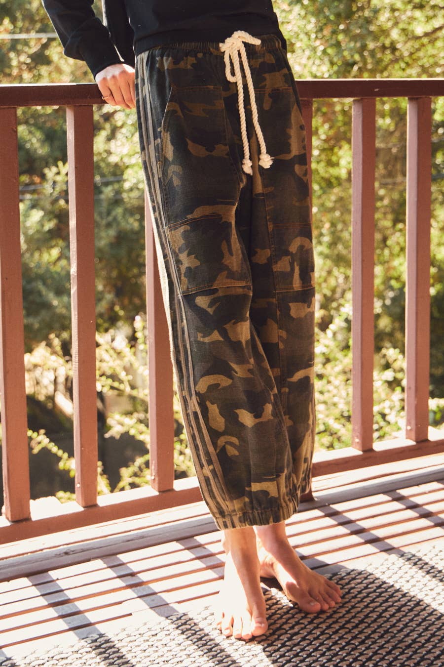 Person wearing camouflage pants standing on a wooden deck with greenery in the background