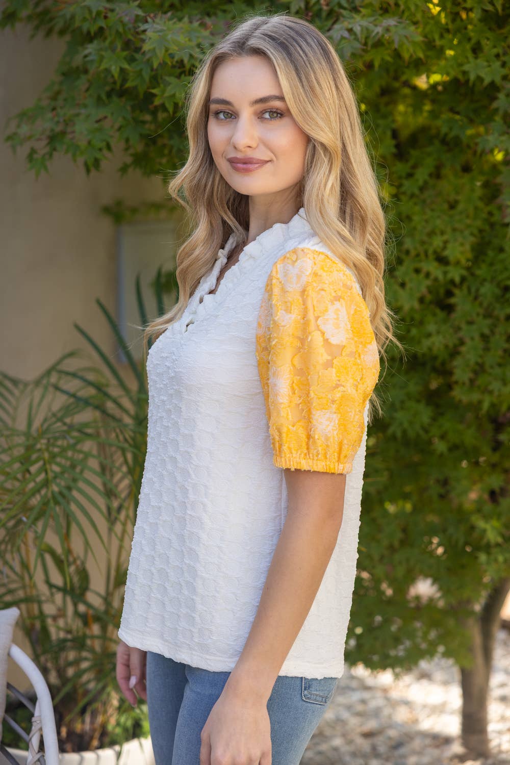Woman wearing a white top with a yellow patterned sleeve against a green outdoor background