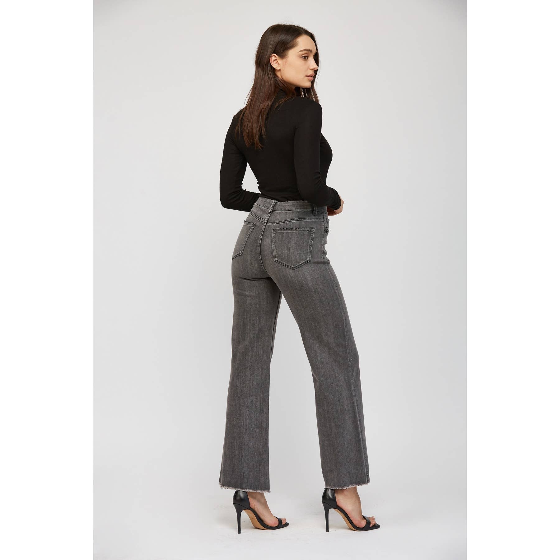 Woman wearing gray high-waisted jeans and a black top with black heels on a white background showing back view.