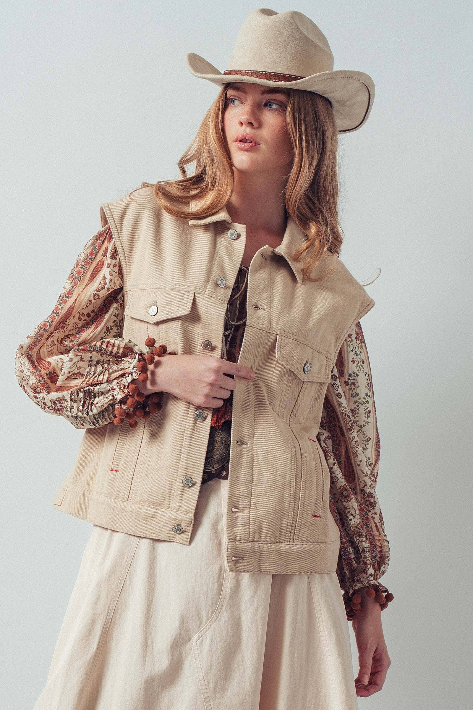 Woman wearing a beige vest, patterned blouse, and cowboy hat against a white background