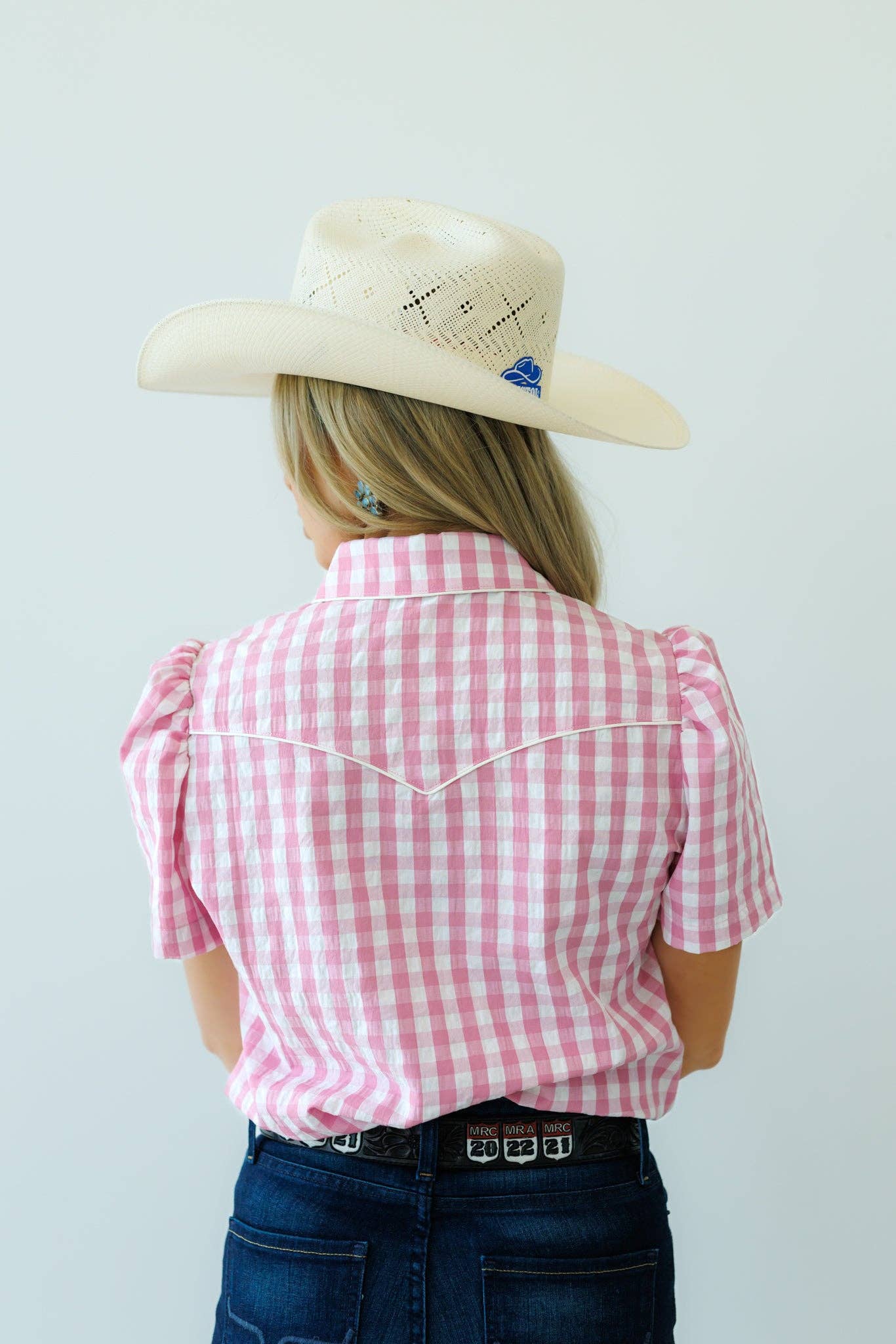 Person wearing a pink checkered shirt and cowboy hat against a white background sowing back view.
